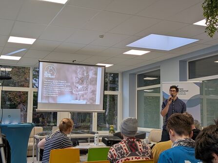 A man holding a microphone, standing infront of a room full of people, with a projector displaying information about the missing lynx project